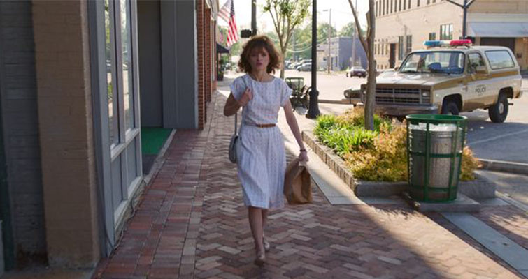 Nancy from Stranger Things walking on a sunny street in the shade of a building.